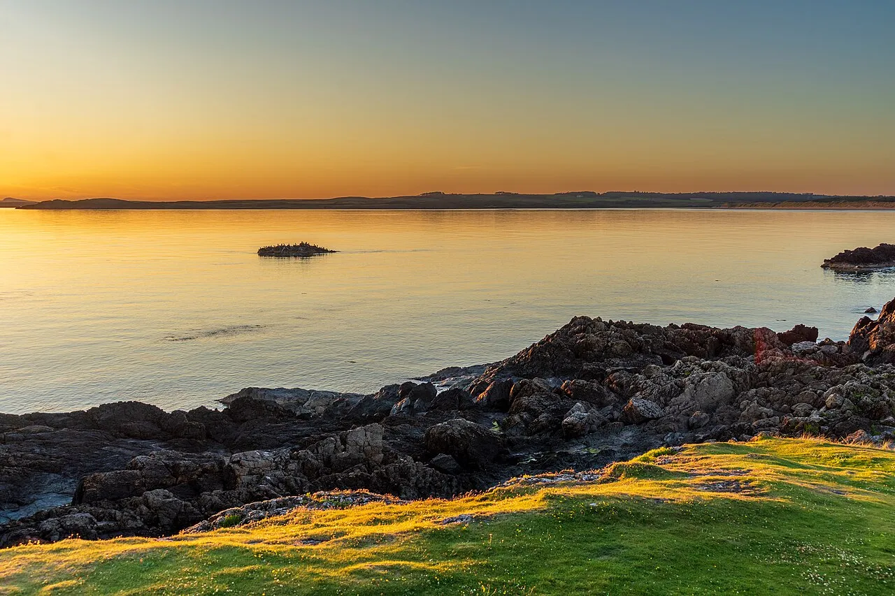 A tidal islet silhouetted in a calm amber sea at sunset, rocky shoreline of Llanddwyn Island in the foreground.