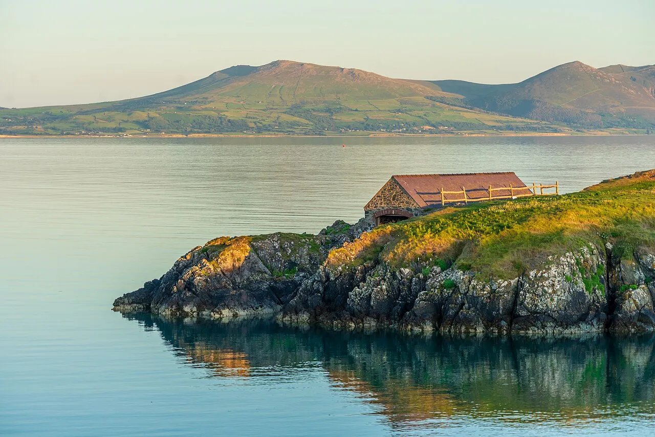 The old stone pilot's boathouse perched on a rocky headland, Snowdonia's mountains reflected in the calm water beyond.