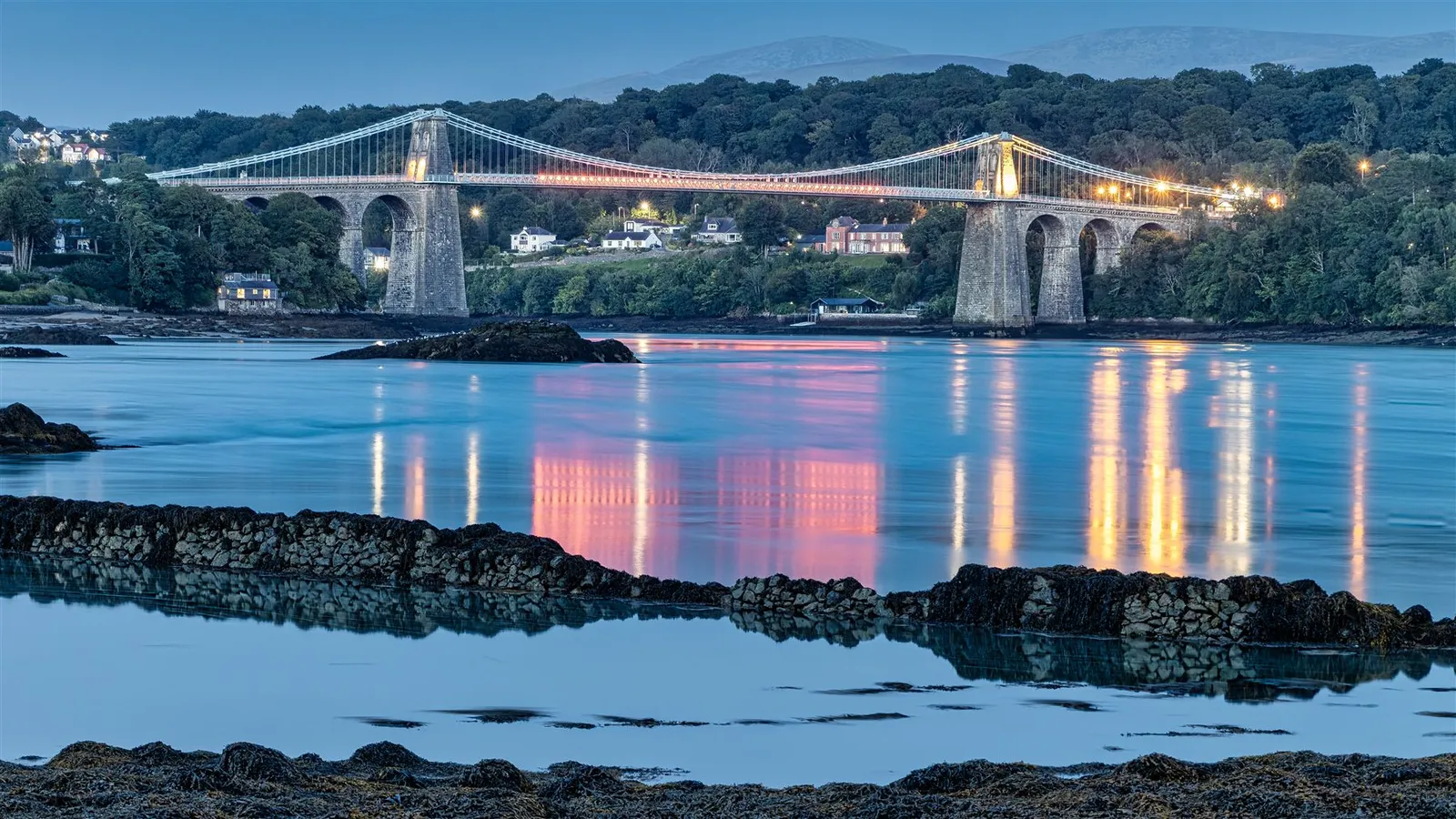 The Menai Suspension Bridge at dusk, its lights and chains mirrored in the still water of the strait below.